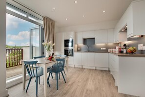 A modern kitchen with white cabinets, a dining area with blue chairs, and a balcony with a view of the countryside.