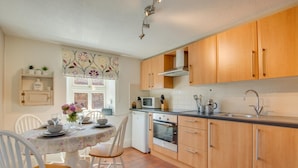 A compact kitchen with wooden cabinets, a built-in oven, and a dining area with a table set for four. - 2 Bed in Sedgeford (oc-2121) (Sedgeford)