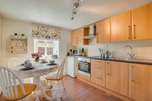A compact kitchen with wooden cabinets, a built-in oven, and a dining area with a table set for four. - 2 Bed in Sedgeford (oc-2121) (Sedgeford)
