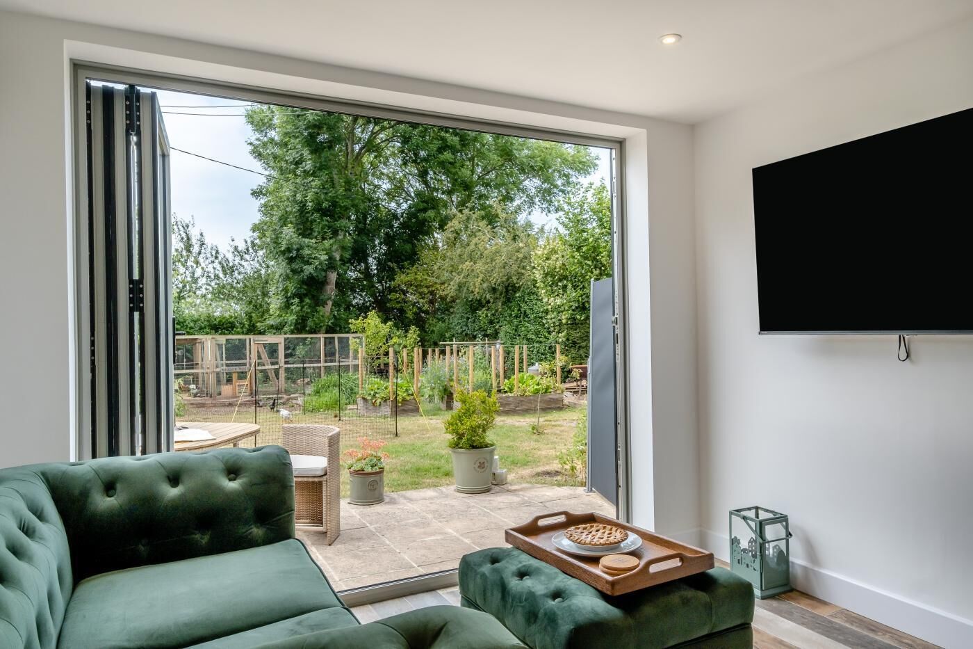 A modern living room with a green sofa, a wooden tray with a decorative item, and a large sliding glass door leading to a garden with a wooden fence and potted plants.