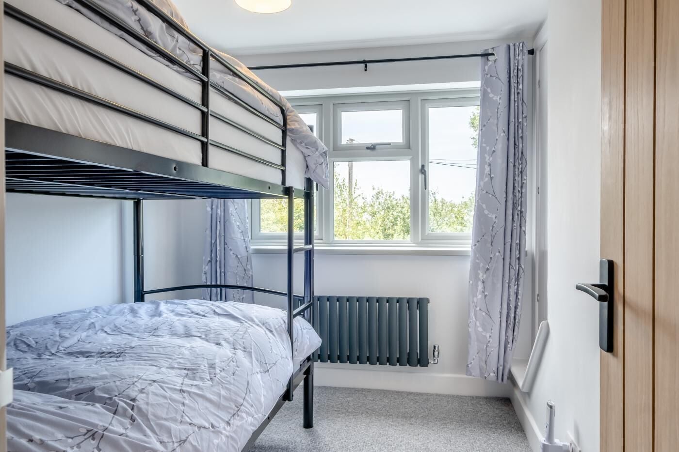 A dormitory room with bunk beds, a window with curtains, a radiator, and a door.