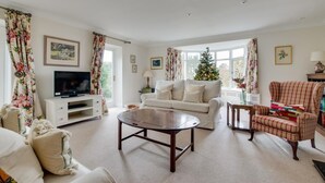 A living room with a white sofa, a wooden coffee table, a television on a white cabinet, a Christmas tree, and floral curtains. - 3 Bed in Cley (oc-2122) (Cley next the Sea)