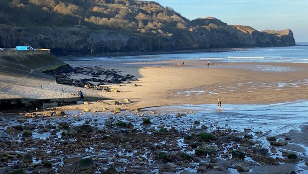 Ein Strand mit felsigem Vordergrund, einem Sandstrand und einer Klippe im Hintergrund.