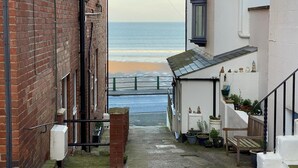 Ein schmaler Gang mit Blick auf den Strand, ein weißes Gebäude mit kleiner Veranda und ein Backsteinhaus.