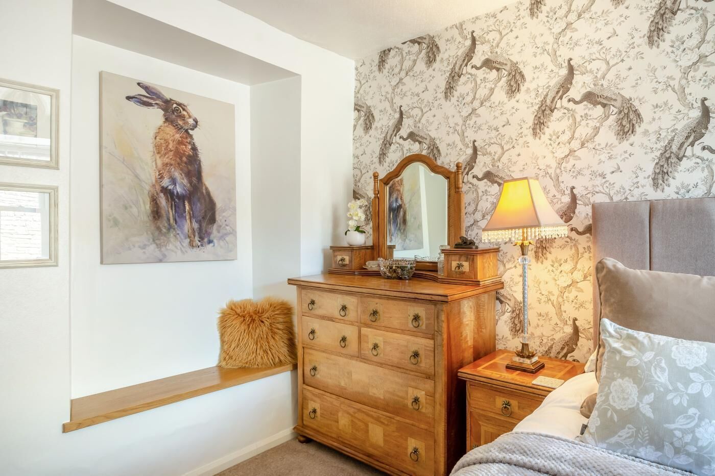 A bedroom with a wooden dresser, a mirror, a lamp, a bed with a floral pillow, and a wall with a peacock wallpaper.