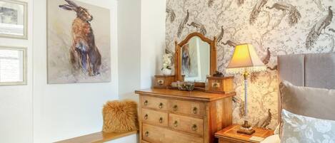A bedroom with a wooden dresser, a mirror, a lamp, a bed with a floral pillow, and a wall with a peacock wallpaper.