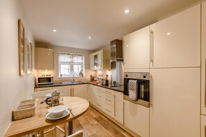 A modern kitchen with a round dining table, built-in appliances, and a window with patterned curtains. - 3 Bed in Whitby (oc-w1003) (Whitby)