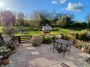 Un espace patio aménagé avec une table et des chaises, entouré de plantes en pot et offrant une vue sur une pelouse verdoyante avec un coin barbecue.