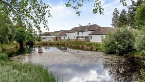 Ein Anwesen am Flussufer mit einem kleinen Teich, umgeben von Grünflächen und einer Reihe weißer Gebäude mit braunen Dächern.