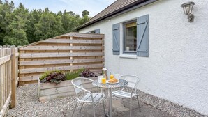 Eine Terrasse mit Frühstückstisch, inklusive Croissants und Saft, neben einem weißen Gebäude mit blauen Fensterläden und einem Holzzaun.