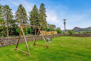 A grassy outdoor area with a wooden swing set, a stone wall, and a bench. - 1 Bed in Newtonmore (oc-l32415) (Newtonmore)