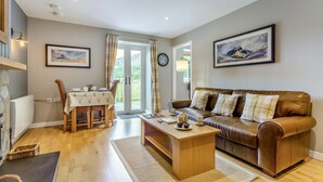 A living room with a brown leather sofa, a wooden coffee table, a dining table with chairs, and a view through a glass door. - 1 Bed in Newtonmore (oc-b32417) (Newtonmore)