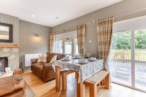 A cozy living room with a fireplace, a dining table set for four, and a large window with curtains. - 2 Bed in Newtonmore (oc-e32418) (Newtonmore)