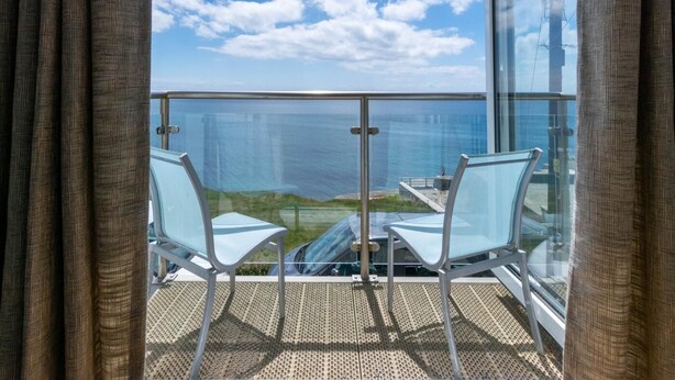En balkon med to stole, gelænder af glas og udsigt over havet.