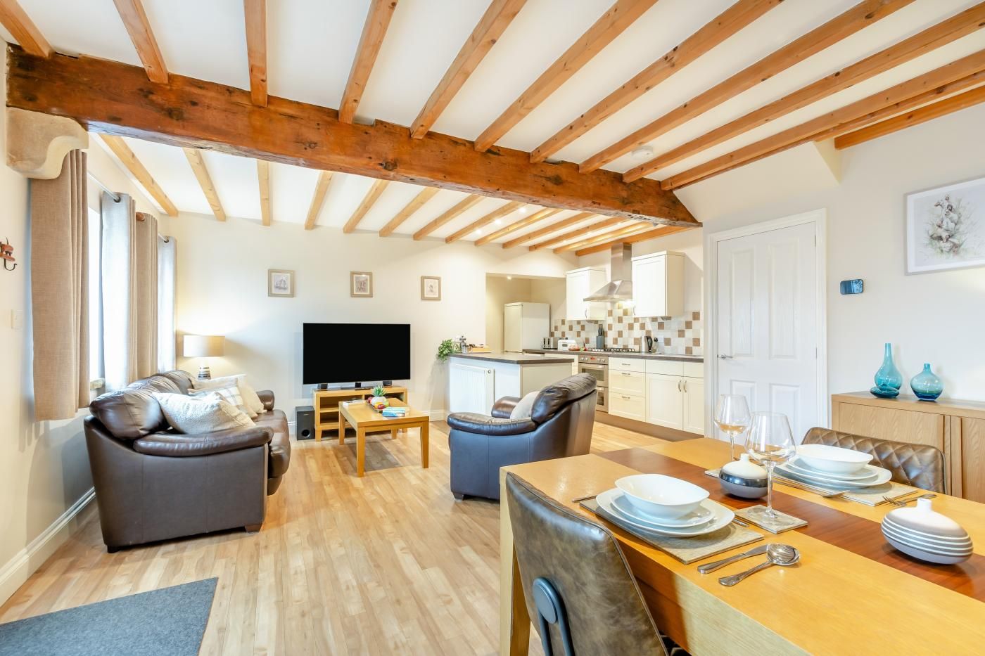 A cozy living room with a wooden ceiling, a flat-screen TV, a sofa, a dining table set for four, and a kitchen in the background.
