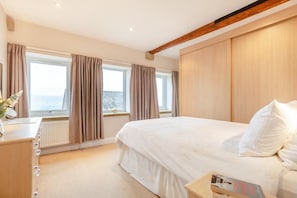 A bedroom with a large bed, a dresser, a window with curtains, and a view of the sea.
