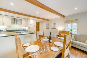 A dining area with a wooden table set for four, a kitchen in the background, and a sofa with patterned curtains. - 3 Bed in Kings Meaburn (oc-cm057) (King's Meaburn)