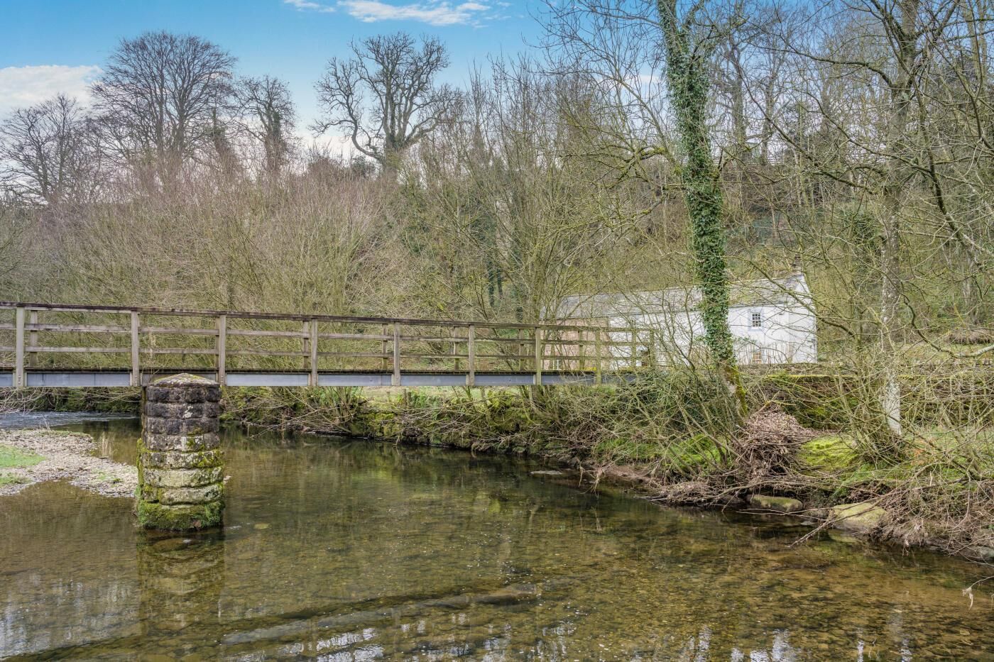 A wooden bridge over a calm river, surrounded by trees and a white building.