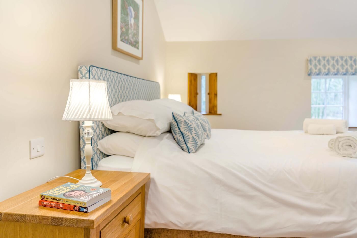 A bedroom with a bed, a bedside table with a lamp, a nightstand, a window with curtains, and a wall-mounted mirror.