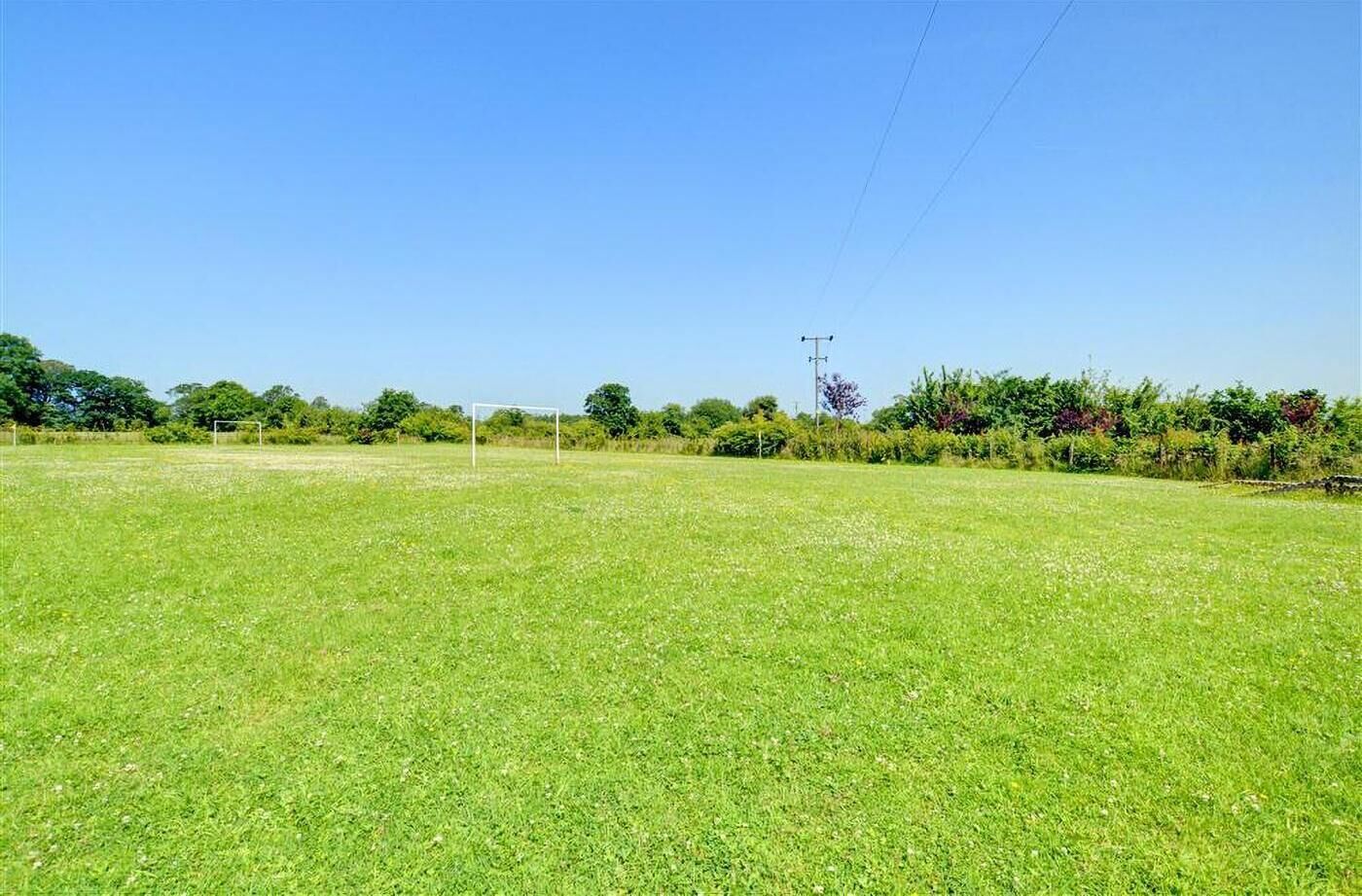 A large, open grassy field with a goalpost in the center, surrounded by trees and a power line in the background.