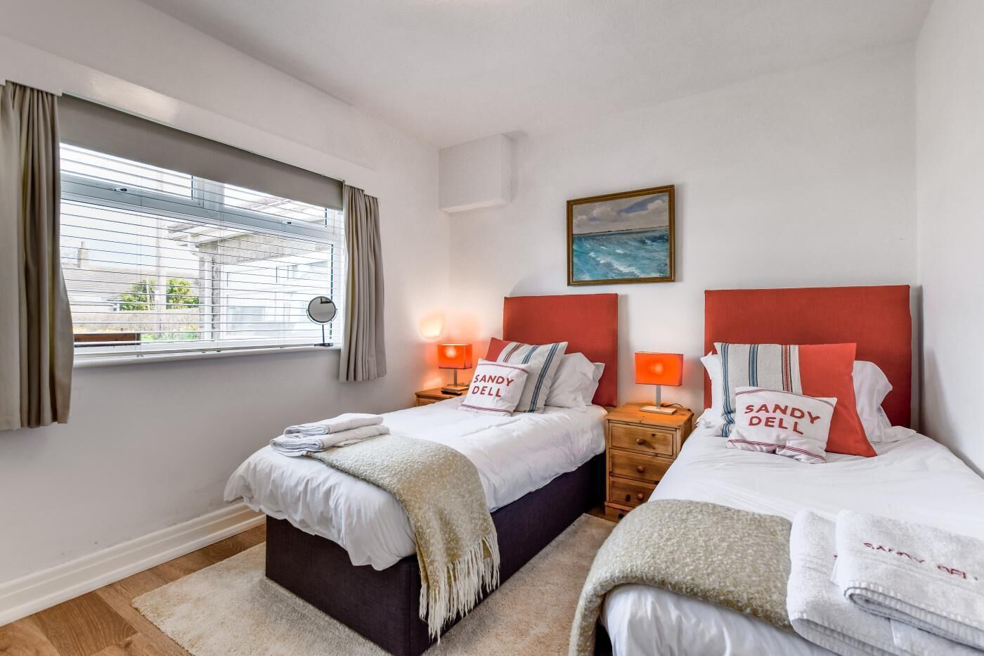 Ein Hotelzimmer mit zwei Betten, jeweils mit Kopfteil, einem Fenster mit Vorhängen, einem Nachttisch mit Lampe und einem Gemälde an der Wand.