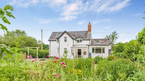 A two-story house with a white exterior, a visible chimney, and a garden with various plants and flowers. - 2 Bed in Yatton (oc-s32603) (Leinthall Earls)