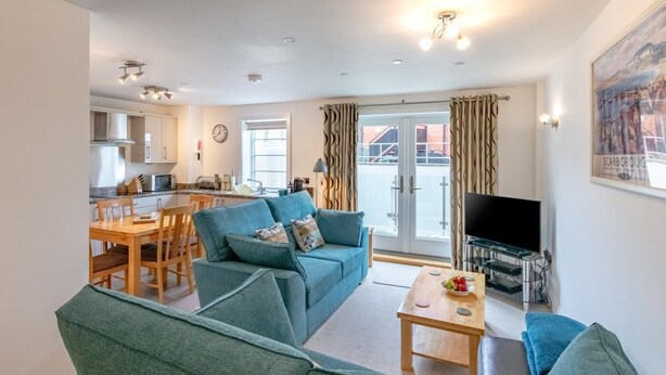 A modern living room with a sofa, a coffee table, and a TV. There is a kitchen area with a dining table and chairs. The room has large windows and a balcony with curtains. - 2 Bed in Whitby (oc-w814) (Whitby)