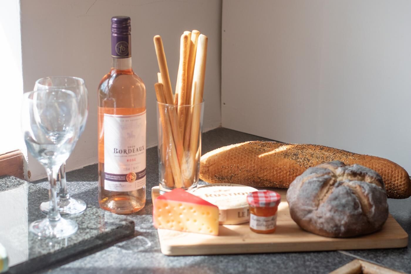 Una mesa con una botella de vino, copas, barras de pan, un pan, queso y un tarro pequeño de mermelada.