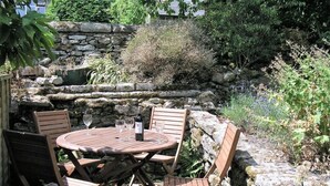 En udendørs terrasse med et rundt træbord og fire stole, omgivet af en stenvæg og forskellige planter.