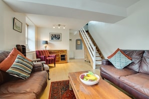 Ein Wohnzimmer mit Ledersofas, einem Holztisch mit Früchten, einem gemusterten Teppich, einer Treppe und einem Essbereich mit einem Schrank.