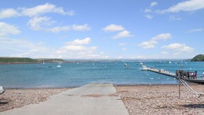 Un paysage côtier avec une plage de galets, une jetée où se trouvent des personnes et des bateaux sur l’eau.