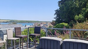 Une terrasse dotée de chaises en osier et d’un espace barbecue, offrant une vue sur un paysage côtier avec des collines et une étendue d’eau.