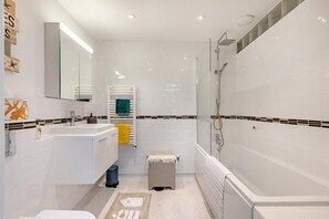 Een moderne badkamer met een wit wastafel, spiegel en douche. Handdoekrek met handdoeken en een prullenbak.