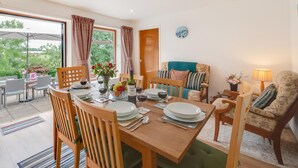 Comedor con una mesa de madera dispuesta para cuatro personas, rodeada de sillas de mimbre. En la mesa hay un florero con flores y una botella de vino. La habitación tiene un ventanal con cortinas, un sofá con cojines y una lámpara sobre una mesita.