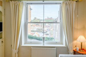A room with a large window, curtains, a radiator, a table with a lamp, and a view of buildings and a castle. - 3 Bed in Whitby (oc-w070) (Whitby)