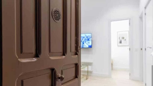 A closed brown door with a decorative panel, leading to a hallway with a white floor and walls, a framed picture on the wall, and a television mounted on the wall. - AR Movistar Arena/Wizink centrally located and modern floor (Madrid)