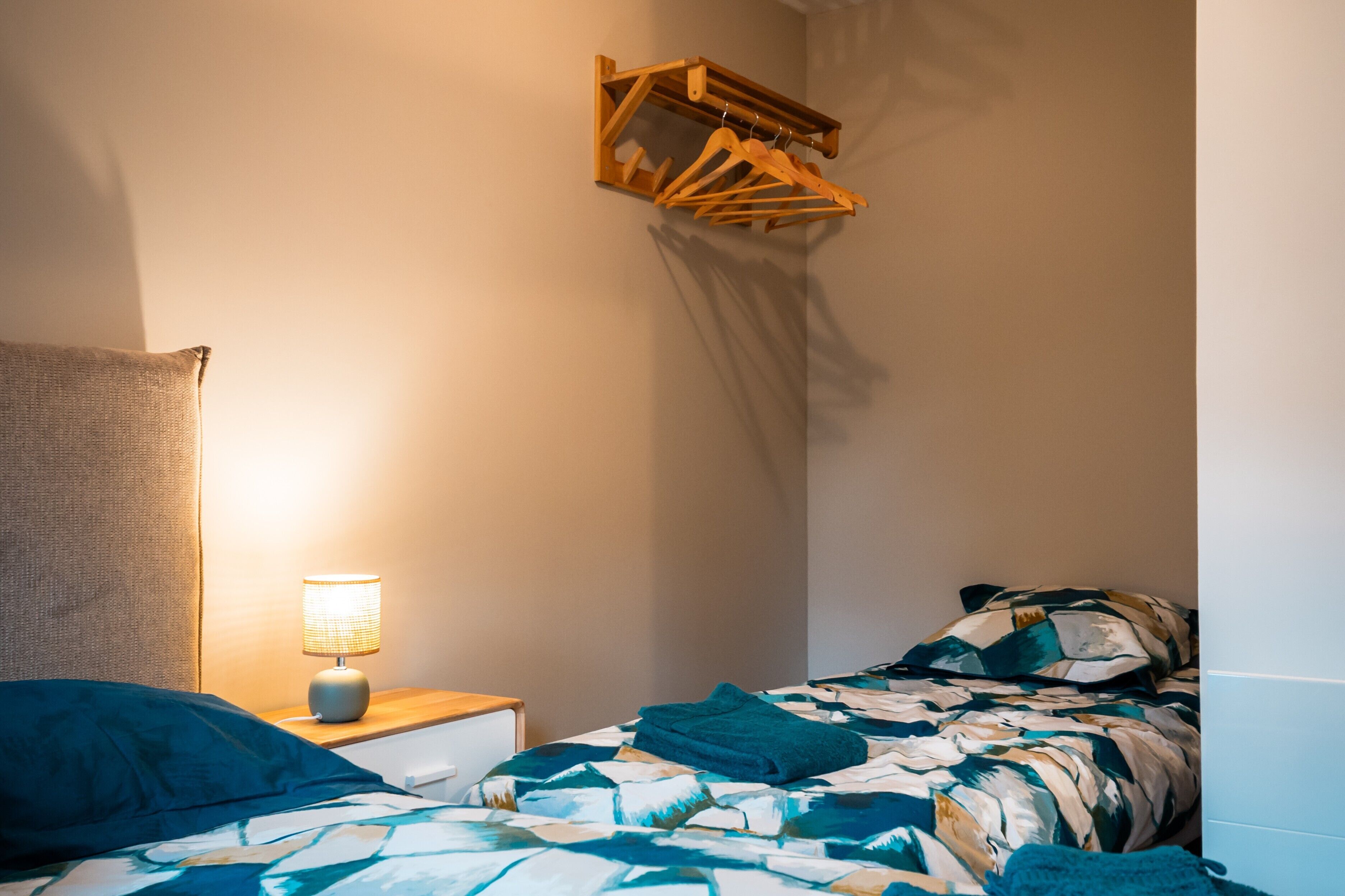 A bedroom with a bed, a nightstand with a lamp, a wooden hanger rack, and a wall-mounted shelf.