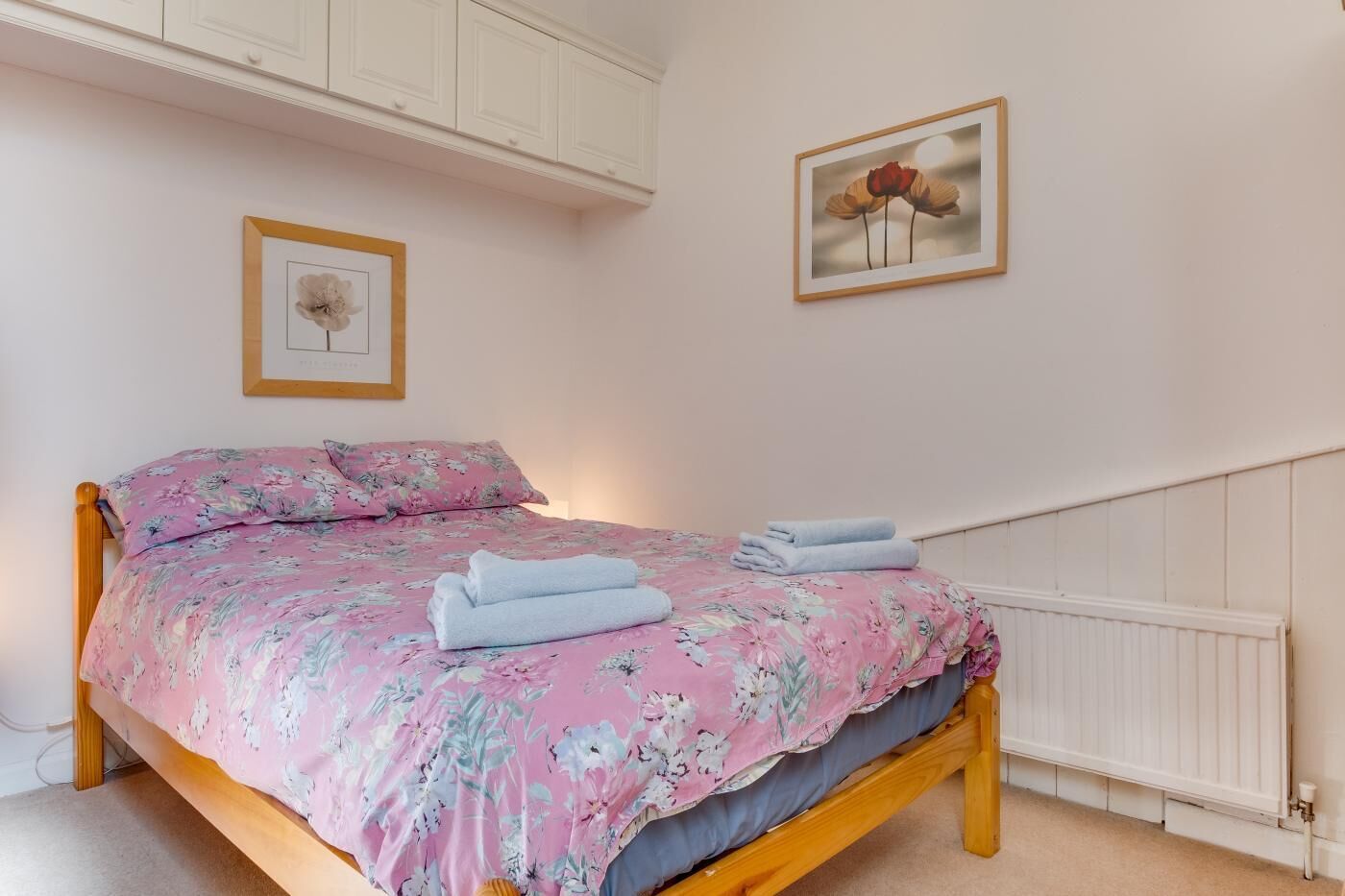 Un dormitorio con cama de madera, ropa de cama floral, un radiador blanco y un cuadro en la pared.