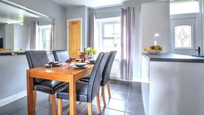 Un espace repas moderne avec une table en bois et des chaises noires, un comptoir de cuisine avec un panier de fruits et une grande fenêtre avec des rideaux.