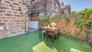 A backyard with a wooden table and chairs, a small garden area with artificial grass, and a brick wall. - 3 Bed in Harrogate (oc-d32616) (Harrogate)