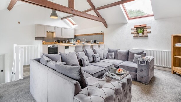 Moderni loft-tyylinen asunto, jossa on esillä olevat puupalkit, suuri harmaa kulmasohva, valkoisilla kaapeilla varustettu keittiö ja kattovalokuvio.