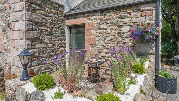 Un edificio de piedra con una puerta, una pequeña mesa con sillas y jardineras con flores moradas.