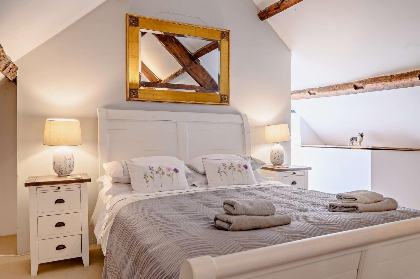 A bedroom with a large bed, two bedside tables, a wooden ceiling beam, and a framed mirror.