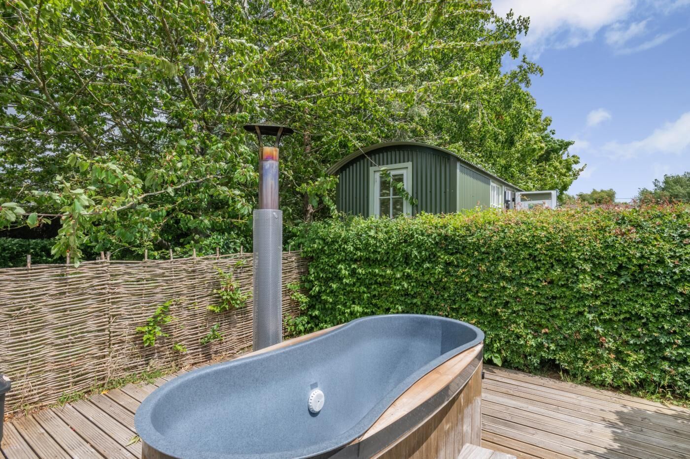 A wooden deck with a hot tub, a metal heater, and a green building in the background.