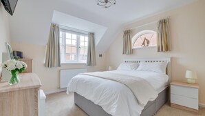 Une chambre avec un grand lit, une table de chevet, une commode, une fenêtre avec des rideaux, un radiateur et un luminaire au plafond.