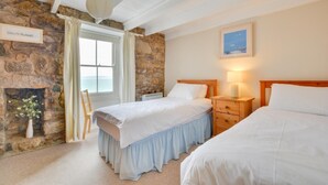 A bedroom with two beds, a stone wall, a window with curtains, a wooden nightstand, and a framed picture on the wall. - 2 Bed in St. Ives (oc-3wd) (St Ives)