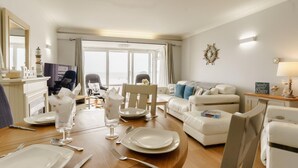Comedor con mesa de madera dispuesta para cuatro personas, un sofá y vista al mar a través de amplios ventanales.