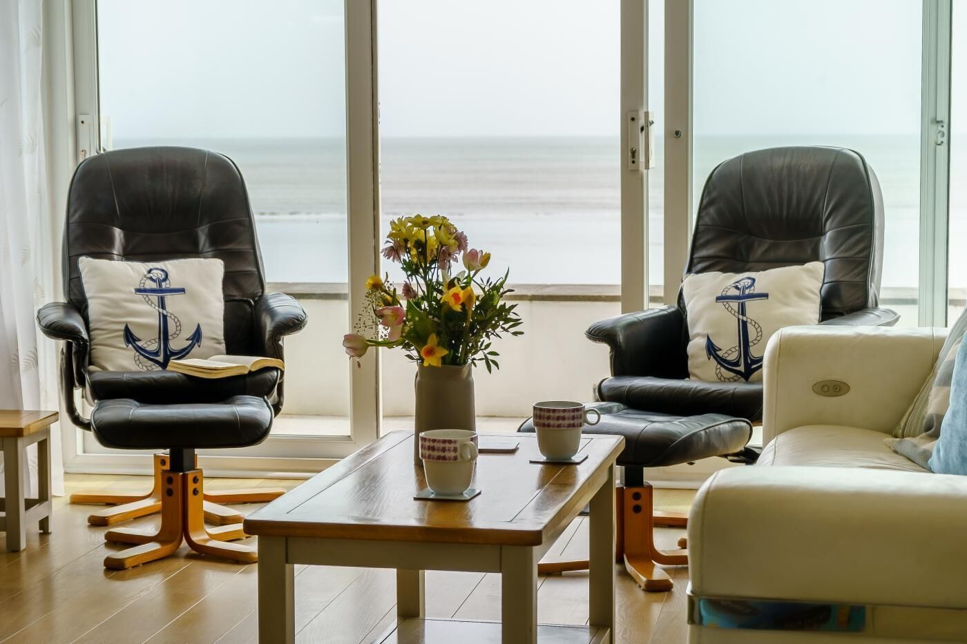 A room with two leather chairs, a small table, a vase with flowers, and a view of the ocean.