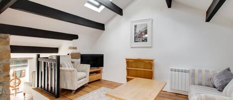 Een loft-woonkamer met zichtbare balken, een houten tafel, een bank, een televisie en een boekenplank.