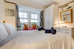 A bedroom with a large bed, a wooden headboard, a window with floral curtains, a white wardrobe, and a mirror with fish decorations. - 3 Bed in Padstow (oc-p00043) (Saint Merryn)